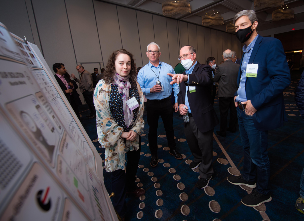 A woman and three men standing by a scientific poster. One of the men points to the poster.