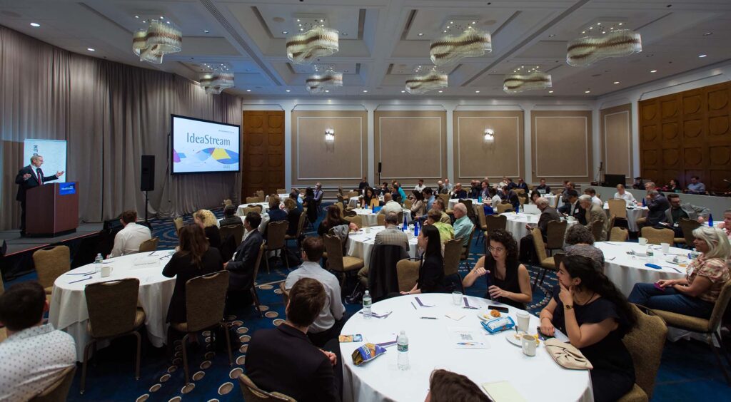 A large ballroom full of people sitting at tables. A man stands at the left making a presentation.